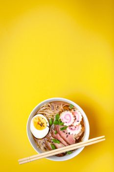 Tasty Japanese noodle soup ramen in white ceramic bowl, meat broth, sliced pork, narutomaki, egg with yolk on pastel yellow background. Traditional dish of Japan, top view, close-up, space for text