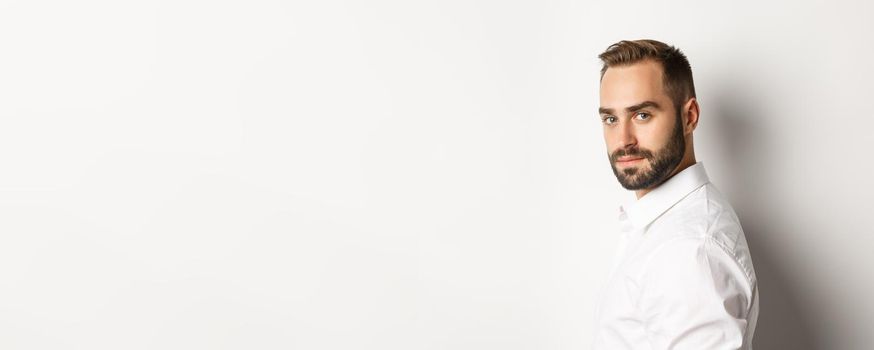 Close-up of professional business man turn face at camera, looking confident, standing against white background.