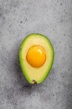 Foods rich in healthy fats for balanced nutrition: raw egg yolk in fresh cut half avocado on gray stone background. Ketogenic low carbs diet or clean eating concept, top view.