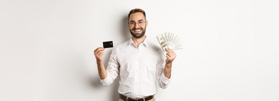 Handsome businessman showing credit card and money dollars, smiling pleased, standing over white background.