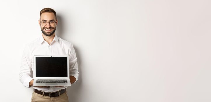 Handsome businessman in glasses, showing laptop screen and smiling happy, standing over white background.