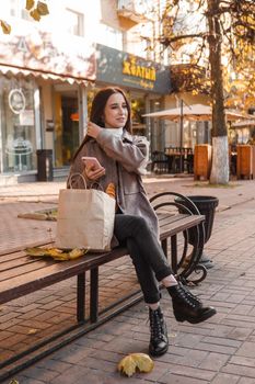A stylish brunette woman walks around the autumn city. The brunette is sitting on a bench outside with a glass of coffee and a phone, holding a craft bag with a baguette.
