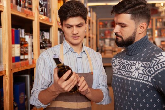 Bearded handsome man shopping for wine at the supermarket, asking sommelier for an advice. Young winemaker working at his shop