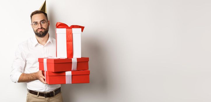 Holidays and celebration. Displeased guy holding birthday gifts and looking disappointed, dislike presents, standing over white background.