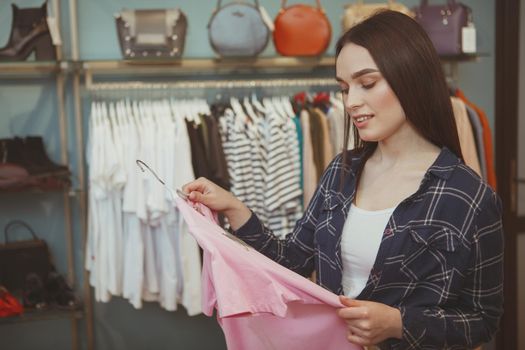 Beautiful young woman examining pink t-shirt on a hanger, enjoying shopping for new clothes, copy space. Attractive relaxed female customer choosing shirt to try at fashion boutique