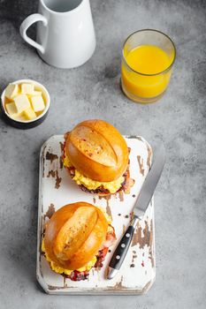 Top view of breakfast sandwiches with scrambled egg, bacon, cheese, tomato on white wooden board, glass with fresh orange juice, white background. Making breakfast concept, selective focus, close up