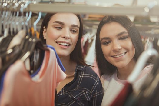 Close up shot of two yougn beautiful women lookign excited, choosing new clothes to buy at fashion boutique. CHeerful female friends enjoying shopping at clothing shop. Retail, buying concept