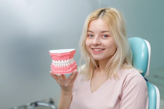 Happy young woman with white healthy teeth holding jaw model sitting in dental chair