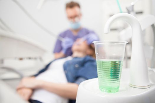 Selective focus on green mouth wash in a cup on dental tap sink, dentist examining teeth of patient on background