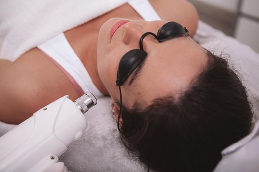 Close up of a woman wearing protective glasses, getting facial hair removed with laser. Young female client relaxing while receiving skin laser treatment