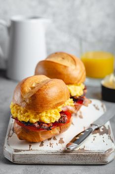 Breakfast sandwiches with scrambled egg, bacon, cheese, tomato on white wooden board, glass with fresh orange juice, white background. Making breakfast concept, selective focus