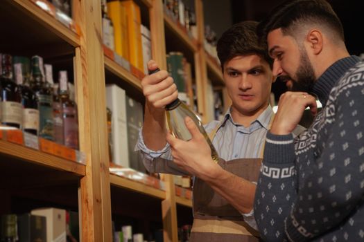 Low angle shot of a professional winemaker helping his customer. Bearded man buying aging wine at the wine store, sommelier helping