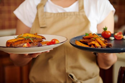 Selective focus on served plates with fried slices of organic batata and roasted sweet potato wedges, drizzled with olive oil and seasoned wit fragrant culinary herbs and rosemary, in chef's hands