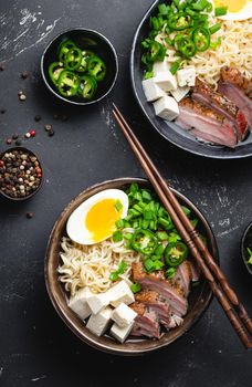 Two bowls of tasty Asian noodle soup ramen with meat broth, tofu, pork, egg with yolk on black rustic concrete background, close up, top view. Hot tasty Japanese ramen soup for dinner asian style