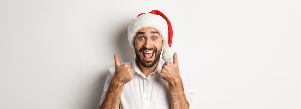 Party, winter holidays and celebration concept. Man enjoying christmas, wearing santa hat and showing thumb up with excited face, white background.