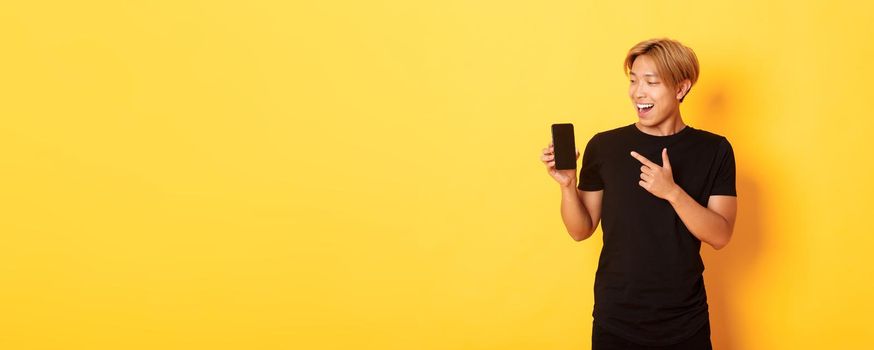 Portrait of satisfied asian guy pointing finger and looking at smartphone screen with pleased smile, showing application, standing yellow background.