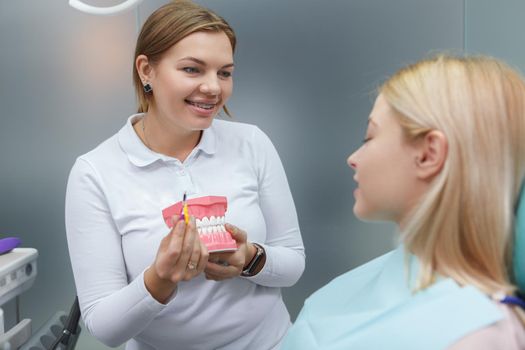 Friendly female dentist with braced teaching her patient correct way of brushing teeth, holding dental model