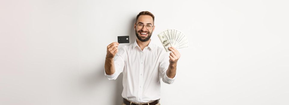 Happy young man showing his credit card and money dollars, smiling satisfied, standing over white background.