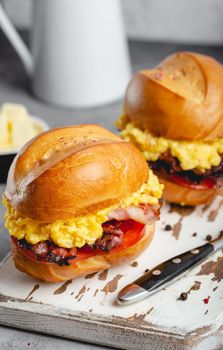 Breakfast sandwiches with scrambled egg, bacon, cheese, tomato on white wooden board, glass with fresh orange juice, white background. Making breakfast concept, selective focus