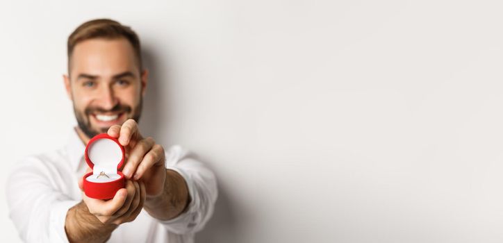 Close-up of handsome man asking to marry him, focus on box with wedding ring, concept of proposal and relationship, white background.