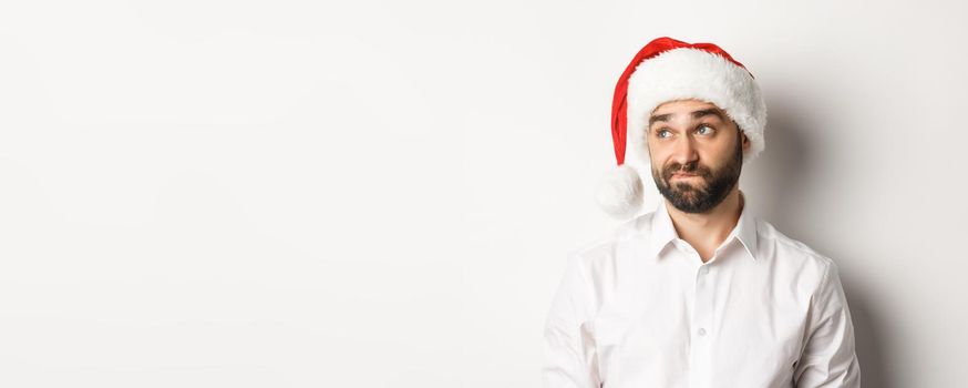 Close-up of skeptical guy in santa hat looking doubtful left, grimacing with hesitation, white background.