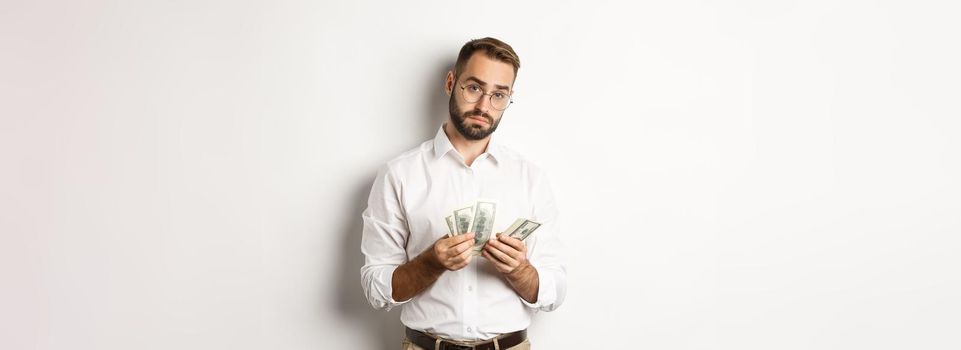 Handsome businessman counting money and looking at camera, standing serious against white background.