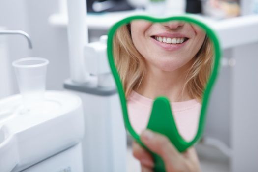 Cropped close up of unrecognizable woman smiling in hand mirror with her healthy white teeth at dental clinic