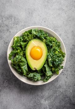 Salad bowl with kale for balanced menu, raw egg yolk in fresh cut half avocado rich in healthy fats on gray stone background. Ketogenic low carbs diet or clean eating concept.