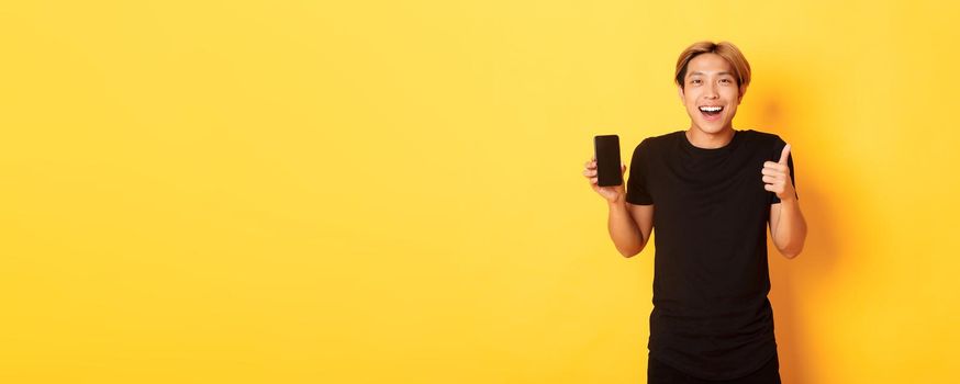 Portrait of pleased smiling asian guy, showing smartphone screen and thumbs-up satisfied with application, standing yellow background.