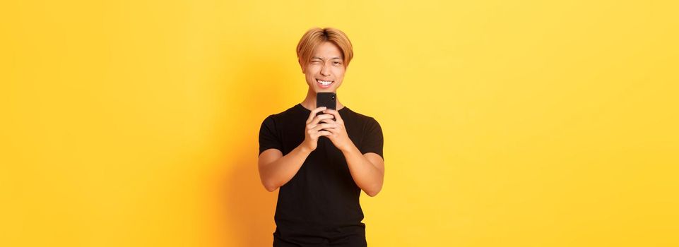 Stylish handsome asian guy taking picture on smartphone and smiling, photographing with mobile phone, standing yellow background.
