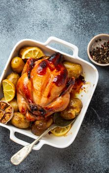 Close-up of baked or roasted whole juicy crispy organic chicken with potatoes, garlic, lemon in white casserole dish with serving spoon on concrete rustic background, top view