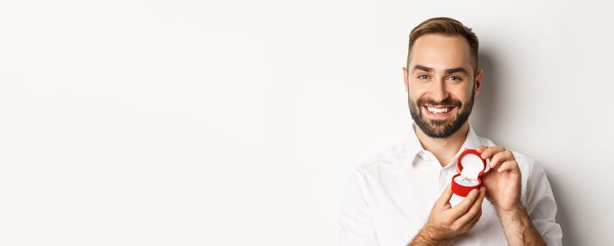 Close-up of happy handsome man making a proposal, holding wedding ring in box and smiling, asking to marry him, white background.