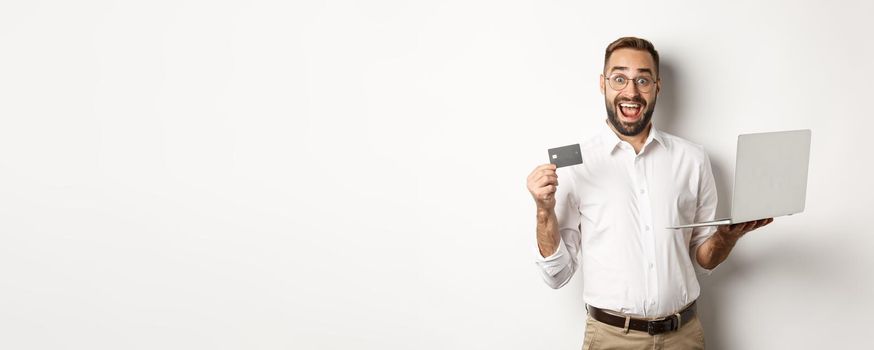 Online shopping. Handsome man showing credit card and using laptop to order in internet, standing over white background.