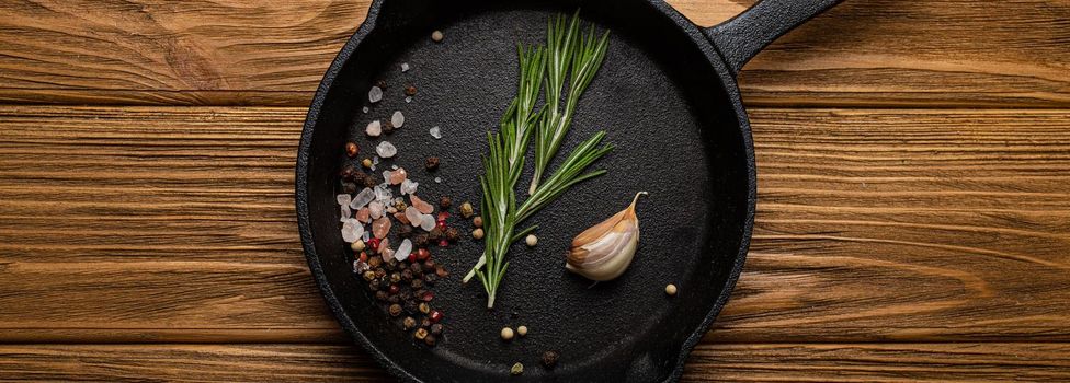 Black cast iron frying pan skillet with food cooking ingredients fresh rosemary, garlic, salt and pepper on rustic wooden table, cooking background and healthy eating kitchen concept