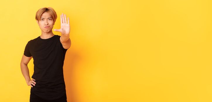Portrait of disappointed serious asian man, smirking displeased and extend hand, showing stop gesture, yellow background.