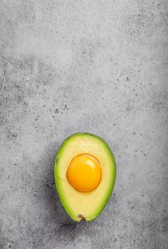 Foods rich in healthy fats for balanced nutrition: raw egg yolk in fresh cut half avocado on gray stone background. Ketogenic low carbs diet or clean eating concept, top view with space for text .