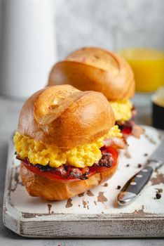 Breakfast sandwiches with scrambled egg, bacon, cheese, tomato on white wooden board, glass with fresh orange juice, white background. Making breakfast concept, selective focus