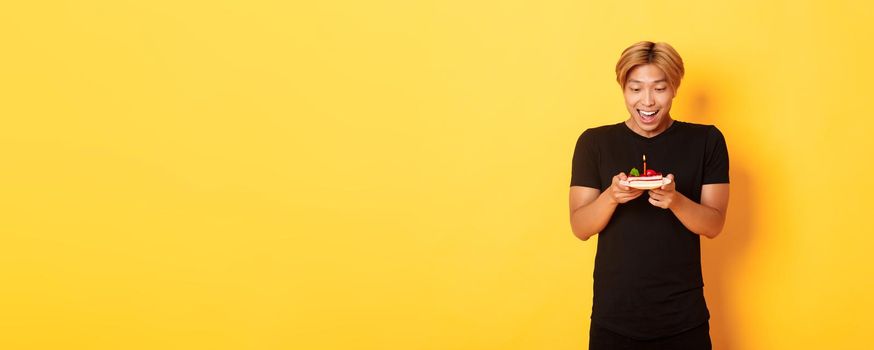 Excited happy attractive asian guy smiling as looking at birthday cake, making wish, celebrating b-day, standing yellow background.