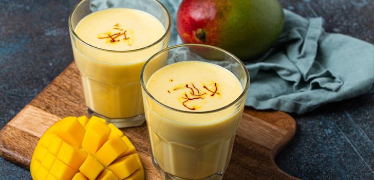 Healthy Indian Ayurveda drink mango lassi in two glasses on rustic concrete table with fresh ripe cut mango, yellow blended beverage made of mango fruit, yoghurt or milk curd and spices