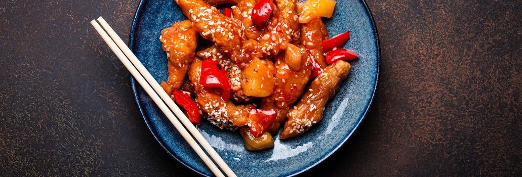 Chinese traditional wok dish sweat and sour deep fried chicken with vegetables stir-fry on plate with sesame seeds on rustic dark concrete stone background top view, Asian meal with chopsticks.