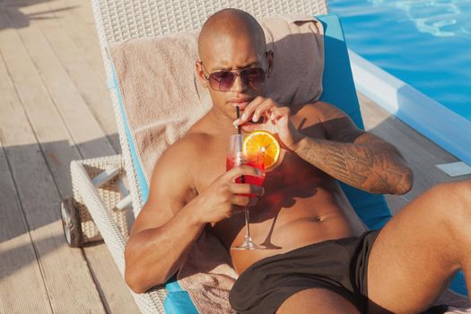 Handsome African man enjoying delicious cocktail at the poolside. Young athletic tattooed man drinking cocktail near the swimming pool while sunbathing