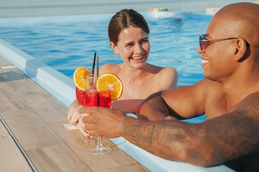 Beautiful happy woman laughing, talking to her boyfriend while having drinks at the swimming pool. Loving multiracial couple celebrating their summer vacation, drinking cocktail at the beach