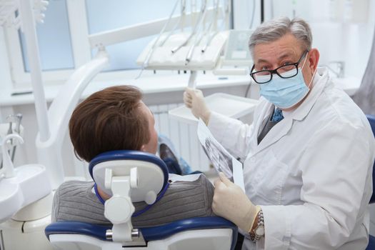 Senior male dentist wearing medical face mask, looking to the camera during dental appointment with patient
