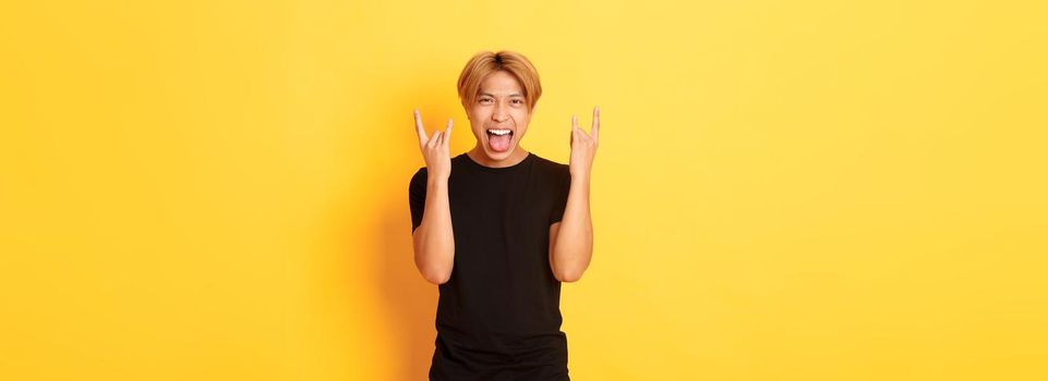 Portrait of joyful handsome asian blond guy having fun, showing rock-n-roll gesture and stick tongue carefree, standing yellow background.