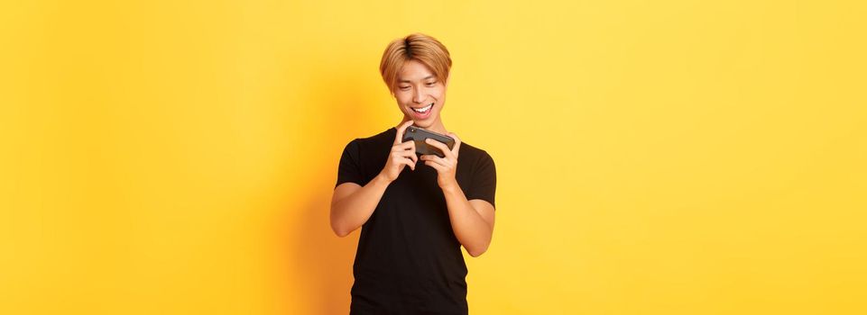 Portrait of stylish handsome asian guy playing smartphone game, looking amused at mobile screen, standing yellow background.