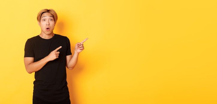 Portrait of curious and amused blond asian guy, pointing fingers upper right corner, showing way, standing yellow background.