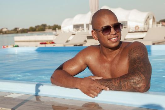 Handsome tattooed African man wearing sunglasses, relaxing at the swimming pool, copy space. Attractive athletic man sunbathing at the poolside, resting after swimming