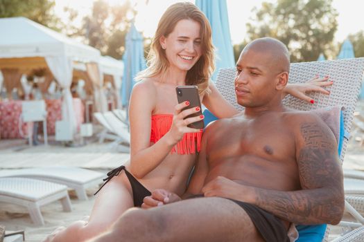 Lovely multiracial couple sunbathing at the beach together, using smart phone. Beautiful woman showing something online to her handsome boyfriend