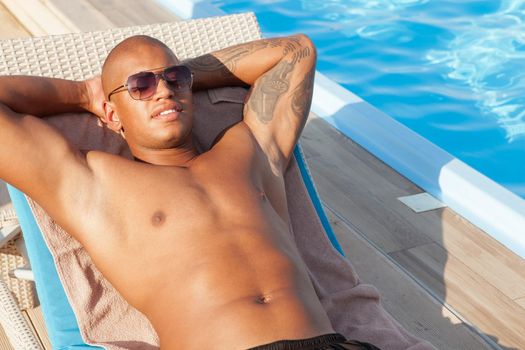 Cropped shot of a handsome African man wearing sunglasses, lying in the sun at the poolside. Attractive man tanning near swimming pool