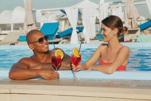 Happy multiethnic couple talking and drinking cocktails in the swimming pool. Charming couple enjoying travelling together, having drinks at the beach